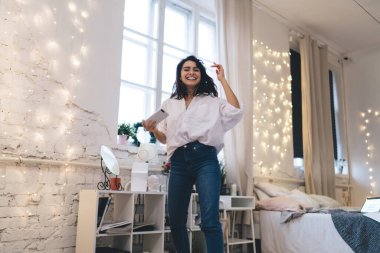 Aşağıdan neşeli, gevşek saçlı ve kollarını kaldırıp kameraya bakarken kulaklıkla müzik dinleyen ve elinde akıllı telefonuyla rahat bir yatak odasına atlayan bir kadın.