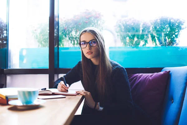 コーヒーを飲みながら木のテーブルに座ってノートを取りながらカメラを見ている長い髪と眼鏡の女性の学生に焦点を当て — ストック写真