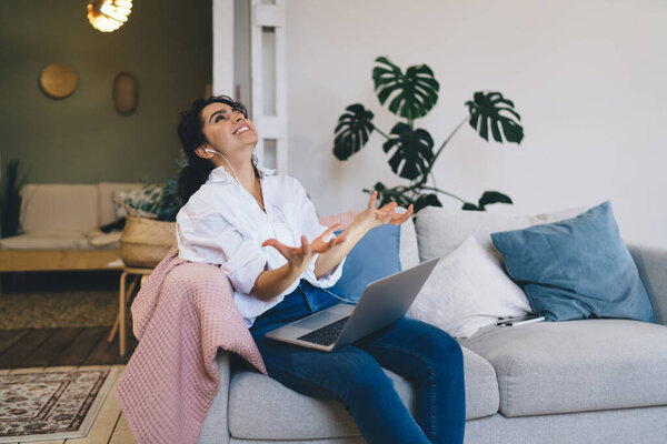 Expressive young female in earphones watching video on netbook and looking up in laughter while resting on comfortable sofa in modern apartment