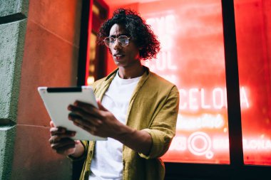 Gözlüklü ciddi yakışıklı hippi elinde dijital tablet tutuyor. 4G internet bağlantısına bağlı. Dalgın giyinmiş erkek, neon ışıklardaki medya içeriğini paylaşmak için touchpad kullanıyor.