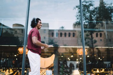 Kentte ellerinde dokunmatik kağıtlarla gezen neşeli turist görüntüsü olumlu ses şarkıları dinlemek için medya uygulamasını kullanıyor. Elektronik kulaklıklı mutlu hipster adam seyahatin keyfini çıkarıyor.