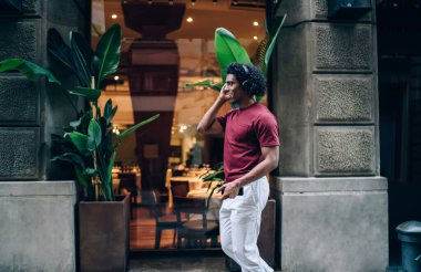 Neşeli erkek turist cep telefonu medya uygulamasını kullanarak şehirde gündüz yürürken pozitif müzik dinledi. Bluetooth kulaklıklı mutlu hipster adam, müzik listesinin tadını çıkarıyor.