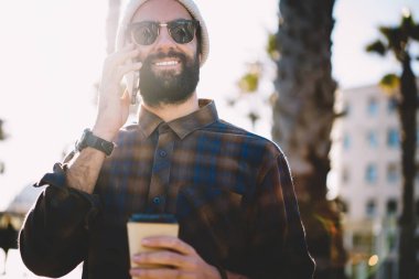 Gezici, güneş gözlüklü, güler yüzlü hippi adamın şehirde cep telefonu kullanarak uluslararası sohbetin tadını çıkararak arkadaşça mobil iletişim kurmak için kahveyle mutlu bir Türk turistin arka planda görüntüsü