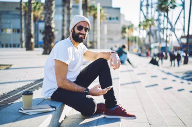 Elinde cep telefonuyla gezen mutlu Türk gezgin portresi, sokak şehirlerinde poz veren, güneş gözlüklü neşeli erkek blogcu kaçış turizmi sırasında 4 g internetli akıllı telefon kullanıyor