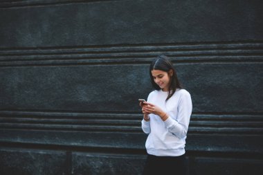 Siyah duvarın yanında beyaz kazak giyen neşeli, genç bir esmer modern cep telefonuyla mesaj atıyor ve gündüz ekrana bakıyor.