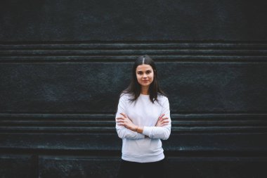 Pozitif, uzun siyah saçlı, beyaz kazaklı, elleri bağlı, taş bina duvarının yanındaki kameraya bakan genç bir kadın.