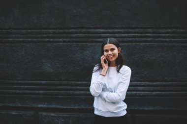 Uzun saçlı, beyaz kazaklı, beton bina duvarının yanında duran ve kameraya bakarken akıllı telefondan iletişim kuran hoş bir genç kadın.