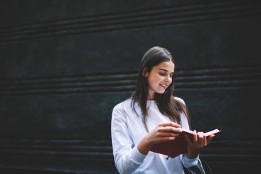 Siyah saçlı, siyah ceketli, siyah taş duvarın yanında durup kırmızı çizgili notları kontrol eden mutlu bir genç kadın.