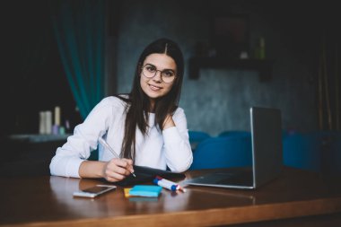 Gündelik kıyafet ve gözlük takan gülümseyen genç bayan ahşap masada otururken akıllı telefon ve dizüstü bilgisayarla not alıyor.