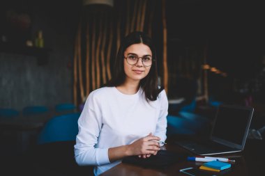 Sıradan giyimli, gözlüklü, sabit dizüstü bilgisayarlı, ahşap masada oturan ve restoranda proje üzerinde çalışırken kameraya bakan hoş bir bayan uzaktan kumanda işçisi.