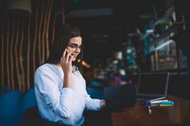 Çalışırken cep telefonuyla konuşurken açık dizüstü bilgisayarı ve not defteriyle masada oturan neşeli kadın çalışanın yan görüntüsü