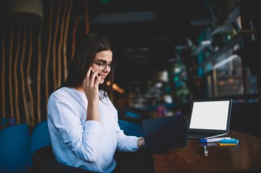 Gülümseyen serbest kadın, beyaz günlük giysiler içinde, gözlüklü, açık dizüstü bilgisayarla masada otururken ve telefonla konuşurken.