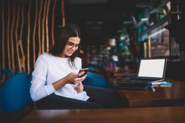 Gülümseyen kadın uzaktan kumandalı günlük giysiler içinde gözlüklü iş sırasında dizüstü bilgisayarla masada oturuyor ve cep telefonuyla sörf yapıyor.