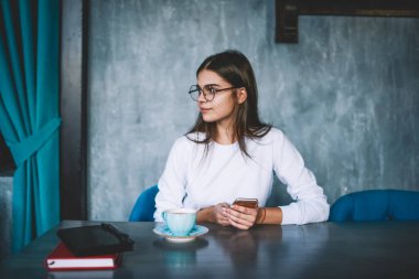 Gündelik giysiler içinde gülümseyen ve gözlüklü genç bir kadın modern kafede bir fincan sıcak içecek içerken cep telefonunu karıştırıyor.