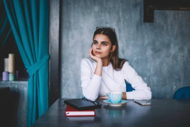 Gündelik giysiler içindeki genç bir kadın bir fincan kahve ve kitapla masada oturuyor ve modern kafeye bakmaktan kaçınıyor.