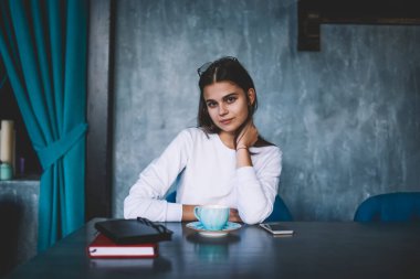 Genç, gülümseyen, beyaz kazak giyen, kahve molası veren, kitaplı, akıllı telefonlu ve kafede bir fincan sıcak içecek içen bir kadın.