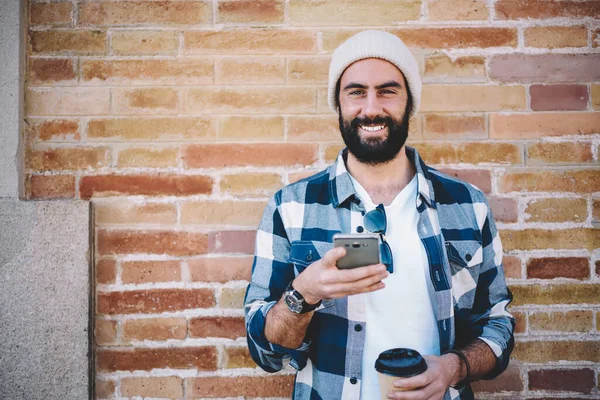 コーヒーと明るい男性ユーザーの半分の長さの肖像画は ネットワークブログのための余暇時間を楽しんで流行の帽子でカメラ 幸せな中東の観光客で笑みを浮かべて手にスマートフォン技術を行く — ストック写真