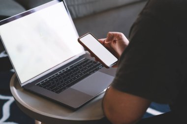 Sıradan gömlekli tanınmayan bir erkek laptopla kahve masasında oturuyor ve evde dinlenirken cep telefonundaki mesajları kontrol ediyor.