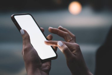 Modern manikürlü, siyah ekranlı modern taşınabilir akıllı telefonlu, esmer bir sokakta bulanık arka planda duran isimsiz, stil sahibi bir kadın.