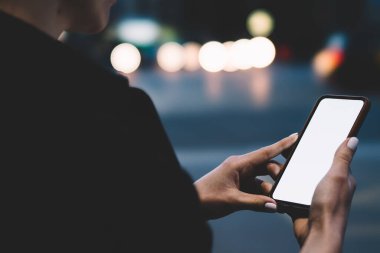 Siyah ceket giyen, cep telefonunu kontrol eden, akşam vakti şehir parkında vakit geçiren kimliği belirsiz bir kadının arka görüntüsü.