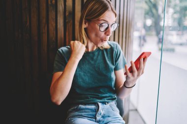 Gündelik giyinen ve gözlüklü neşeli bayan cam pencerenin yanındaki ahşap arka planda modern çalışma alanında mesajlaşırken neşeli bir jest ile akıllı telefon ekranına bakıyor.