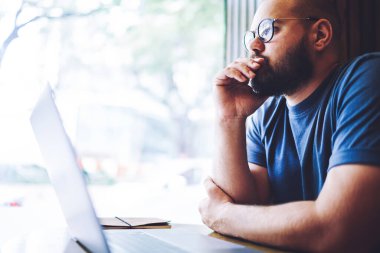 İş günü açık dizüstü bilgisayarla masada otururken dudaklarına dokunan tişörtlü erkek işçinin yan görüntüsü