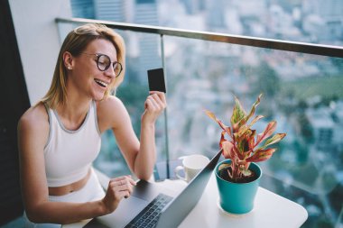 Spor kıyafetli yüksek açılı genç bir kadın evde dizüstü bilgisayarla alışveriş yapıyor ve iyi bir indirim aldığına seviniyor.