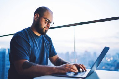 Ciddi sakallı, sıradan kıyafetleri olan bir adamdan. Masada oturmuş dizüstü bilgisayara bakıyor ve proje için bilgi topluyor.