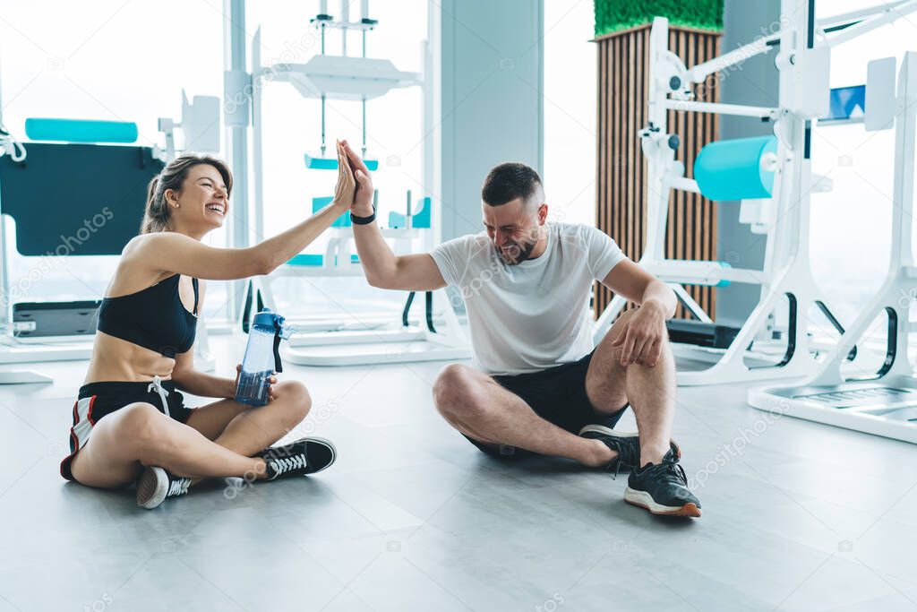 Full body of laughing fit couple giving five while sitting on floor and having break during workout in modern gym