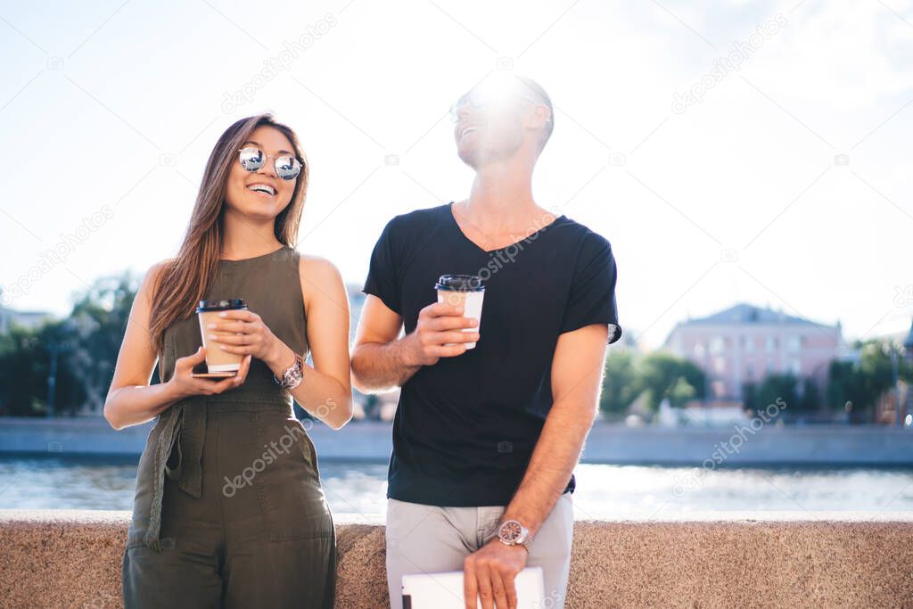 Backlit view of cheerful hipster guys with disposable takeaway cups enjoying friendly meeting during sunny day discussing relationship and smiling, diverse couple in love talking during coffee break