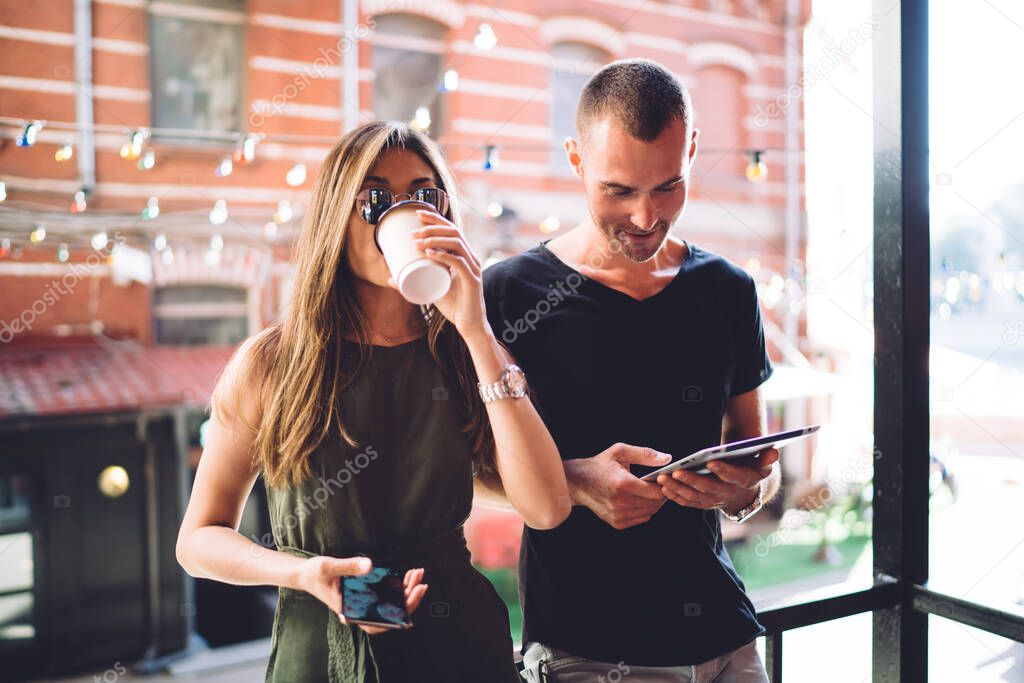 Half length of casual dressed hipster girl with modern mobile phone drinking tasty caffeine beverage while Caucasian boyfriend browsing web publication on digital tablet, millennial persons