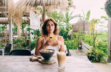 Cep telefonuyla kafeterya masasında kahvaltı kasesi ve latte macchiato ile kameraya bakan genç, tatminsiz etnik kadın.