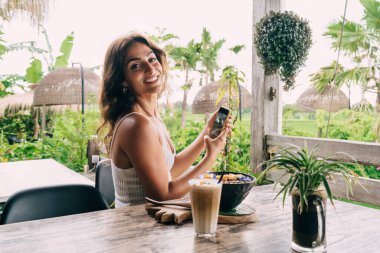 Yan görünüşlü mutlu genç bayan cep telefonuyla geziyor ve tropikal bahçede lezzetli egzotik yiyeceklerle masada oturuyor kameraya bakıyor ve gülümsüyor.