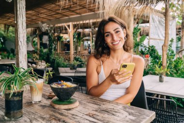 Kafeterya masasında cep telefonuyla lezzetli yemekler ve kahveyle mutlu bir etnik bayan yazın terasta kameraya bakıyor.