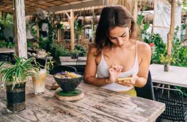 Egzotik kafeteryadaki ahşap masada yemek yerken akıllı telefondan mesaj okuyan şık, genç bir etnik kadına odaklanmış.