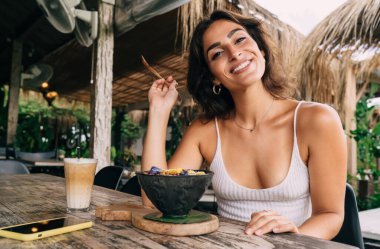 Elinde kahvaltı kasesi ve akıllı telefonun yanında bir bardak kahveyle oturmuş kameraya bakarken dirseğinde kaşık olan pozitif etnik bir kadın.