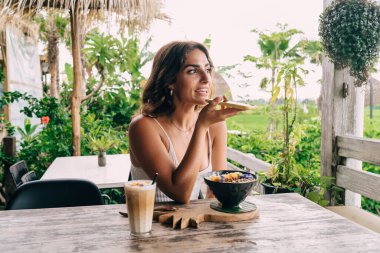 Neşeli etnik bayan, tropik tatil beldesindeki kafeteryada kahvaltı kasesi ve kahveyle masada otururken cep telefonundan sesli mesaj kaydediyor.