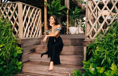 Tam vücut pozitifliği olan çıplak ayaklı kadın şık giysiler içinde caddelerde dinleniyor. Etrafı bitkilerle çevrili ahşap bir verandada güneşli bir günde yaz kafenin iç kısımlarını süsleyen saksılara karşı oturuyor.