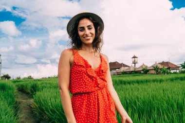 Kıvırcık saçlı, şık kırmızı elbiseli ve şapkalı, kameraya bakıp pirinç tarlasında gülümseyen genç, pozitif etnik bir kadın.