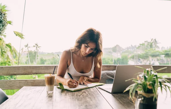 Concentrated Young Ethnic Businesswoman Taking Notes Copybook While Sitting Table — Stock fotografie