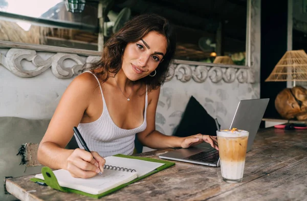 若い幸せな民族の女性のリモート従業員は コーヒーとテーブルでネット上に入力しながら 日記に書いてカメラを見て — ストック写真