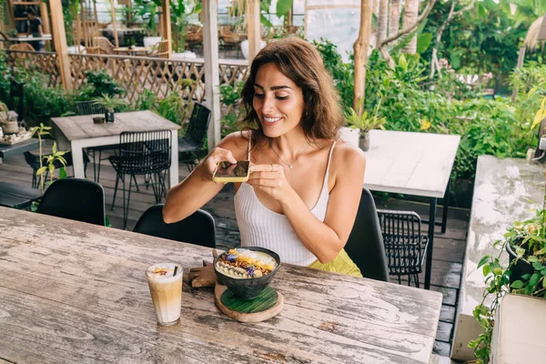 Genç, etnik bir kadın yazın terastaki kafe masasında dinlenirken cep telefonuyla iştah açıcı kahvaltı kasesinin fotoğrafını çekiyor.