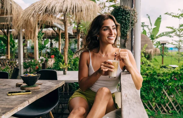 夏の日を眺めながらカフェに座ってカクテルを飲みながらテーブルに座っているカジュアルな服の正の民族の女性 — ストック写真