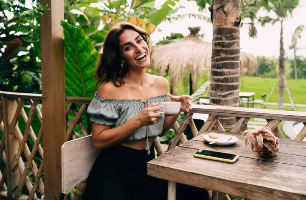トロピカルガーデンのカフェのベランダに座って朝食中にコーヒーを飲むスタイリッシュな夏の服で楽しい若い民族の女性 — ストック写真