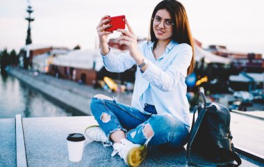 Optik gözlüklü neşeli hippi kızın portresi kameraya gülümserken cep telefonu uygulamasını kullanarak vlog içeriğini etkiliyor ve seyahat sitesine paylaşıyor, mutlu Kafkasyalı blogcu