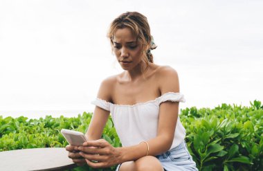 Hafta sonu tropikal tatil beldesindeki otelin terasında dinlenirken cep telefonuyla mesajlaşan yaz kıyafetleri içindeki ciddi genç bayan.