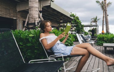 Yazlık kıyafetlerle, sandalyede oturup kitap okuyan ve tropik tatil beldelerinin terasında vakit geçiren çıplak ayaklı çekici bir kadın.