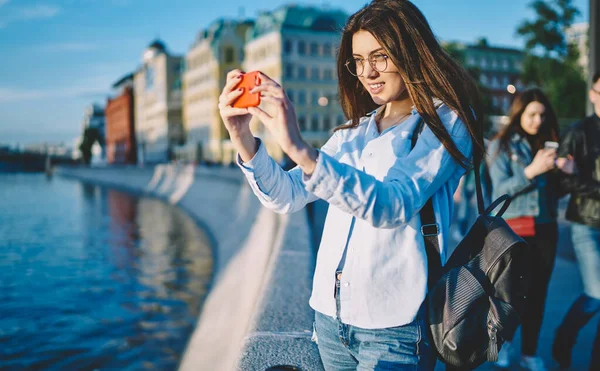 Gözlüklü beyaz kadın etkileyicisi cep telefonu aygıtı kullanarak göz düzeltmesi sağlıyor. Topraktaki manzara resimlerini tıklıyor. Milenyum turisti sırt çantasıyla gezici video çekiyor.