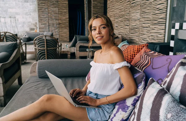 Modern otelin avlusunda dizlerinin üzerine çöküp renkli yastıklarla rahat koltukta oturan ve klavyede daktilo kullanan modaya uygun giyinmiş genç bir kadın.