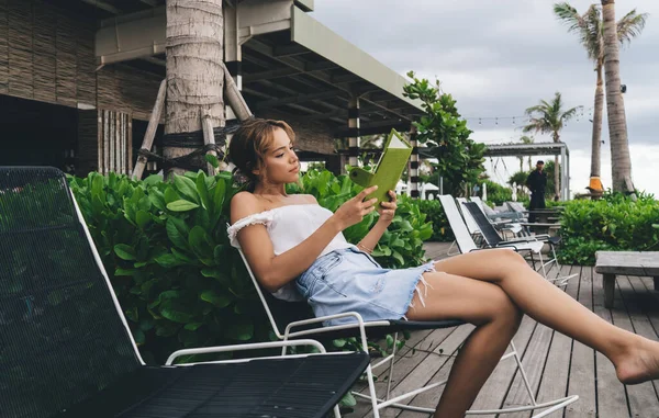 Yazlık kıyafetlerle, sandalyede oturup kitap okuyan ve tropik tatil beldelerinin terasında vakit geçiren çıplak ayaklı çekici bir kadın.